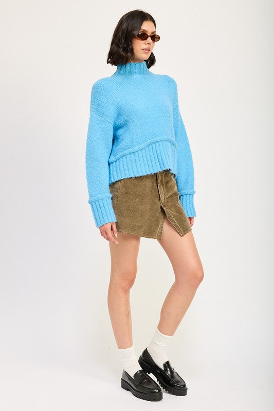 A woman wearing a light blue Turtle Neck Body Sweater, brown sunglasses, a brown crossbody bag, and a khaki skirt stands against a plain background.