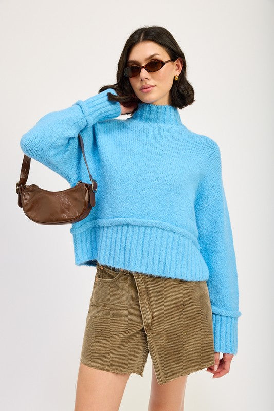 A woman wearing a light blue Turtle Neck Body Sweater, brown sunglasses, a brown crossbody bag, and a khaki skirt stands against a plain background.