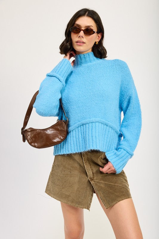 A woman wearing a light blue Turtle Neck Body Sweater, brown sunglasses, a brown crossbody bag, and a khaki skirt stands against a plain background.