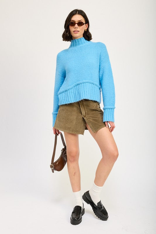 A woman wearing a light blue Turtle Neck Body Sweater, brown sunglasses, a brown crossbody bag, and a khaki skirt stands against a plain background.