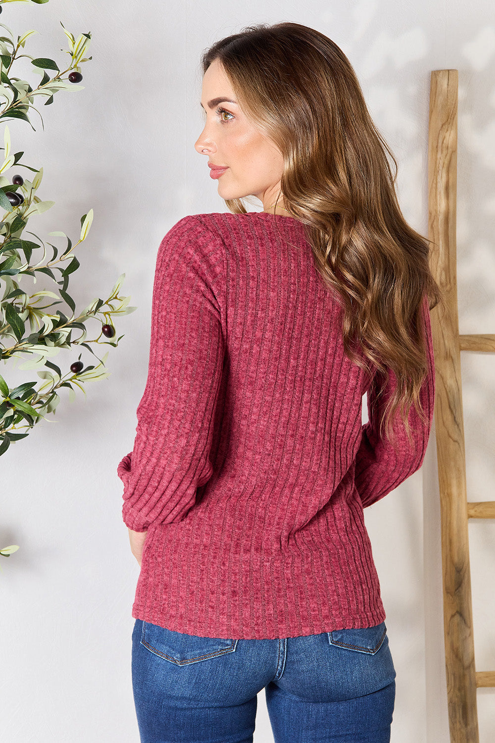 A person in a stylish maroon Ribbed Round Neck Lantern Sleeve Blouse and jeans stands by a wooden ladder, with olive branches in the background.