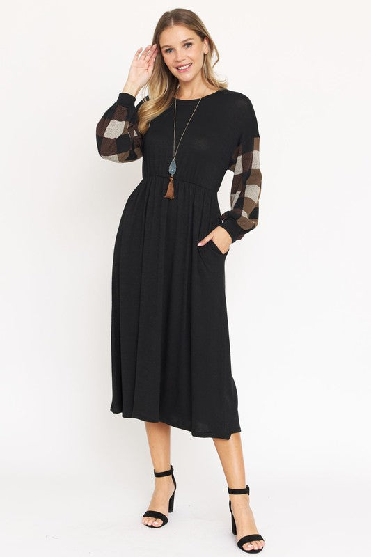 A woman stands against a plain white background wearing the Knit Bishop Sleeve Tea Length Dress, featuring a black tea length design with plaid knit bishop sleeves. She has her hands in the dress pockets and is wearing black heels.