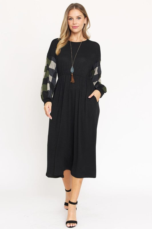 A woman stands against a plain white background wearing the Knit Bishop Sleeve Tea Length Dress, featuring a black tea length design with plaid knit bishop sleeves. She has her hands in the dress pockets and is wearing black heels.