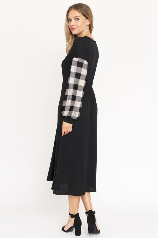 A woman stands against a plain white background wearing the Knit Bishop Sleeve Tea Length Dress, featuring a black tea length design with plaid knit bishop sleeves. She has her hands in the dress pockets and is wearing black heels.