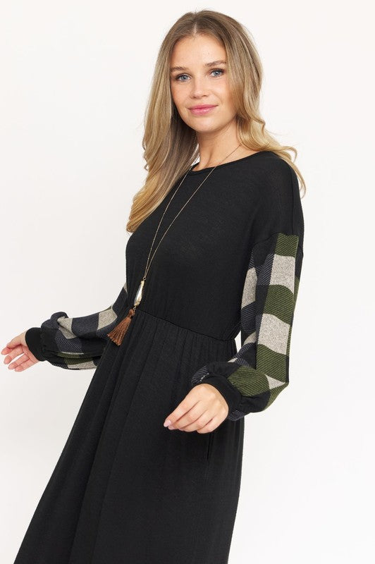 A woman stands against a plain white background wearing the Knit Bishop Sleeve Tea Length Dress, featuring a black tea length design with plaid knit bishop sleeves. She has her hands in the dress pockets and is wearing black heels.