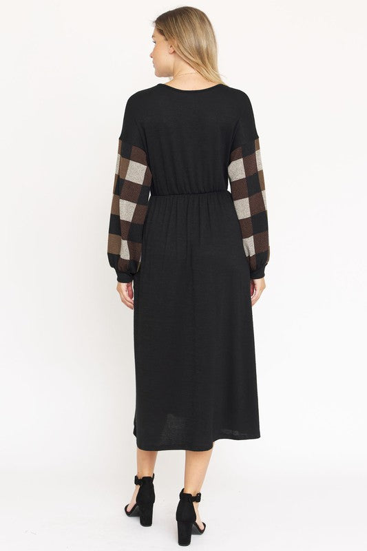 A woman stands against a plain white background wearing the Knit Bishop Sleeve Tea Length Dress, featuring a black tea length design with plaid knit bishop sleeves. She has her hands in the dress pockets and is wearing black heels.