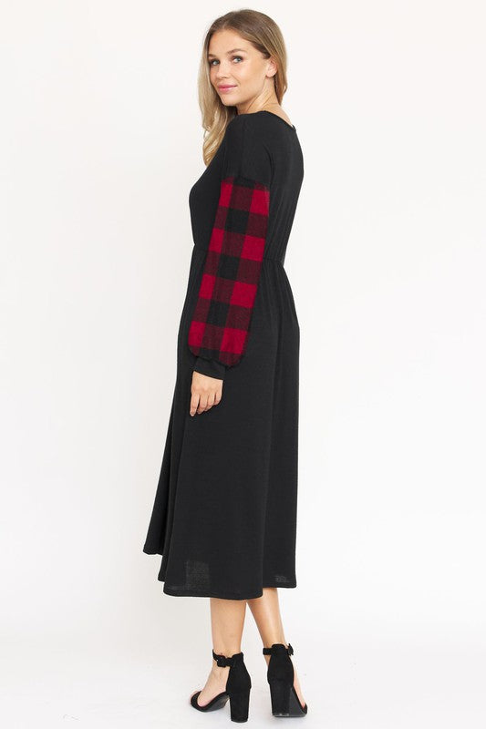 A woman stands against a plain white background wearing the Knit Bishop Sleeve Tea Length Dress, featuring a black tea length design with plaid knit bishop sleeves. She has her hands in the dress pockets and is wearing black heels.