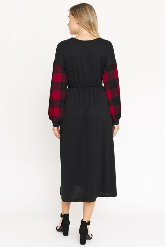 A woman stands against a plain white background wearing the Knit Bishop Sleeve Tea Length Dress, featuring a black tea length design with plaid knit bishop sleeves. She has her hands in the dress pockets and is wearing black heels.