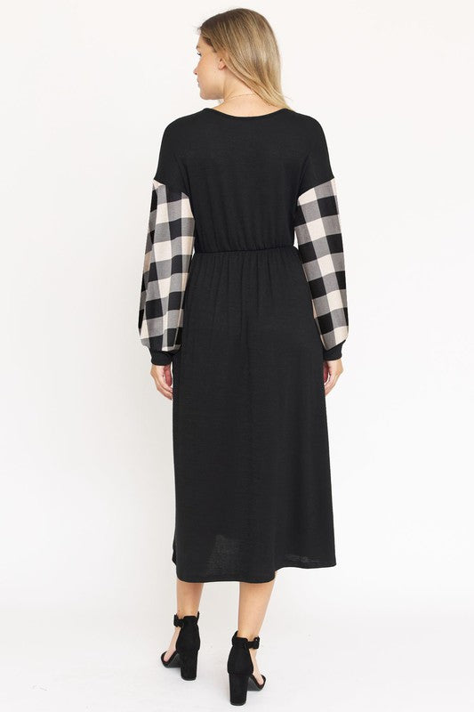 A woman stands against a plain white background wearing the Knit Bishop Sleeve Tea Length Dress, featuring a black tea length design with plaid knit bishop sleeves. She has her hands in the dress pockets and is wearing black heels.