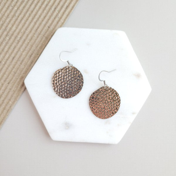 The Lucia Earrings - Silver feature round textured metal designs with stainless steel hooks, arranged on a white hexagonal marble surface at a diagonal angle, set against a subtle beige background. These elegant accessories are crafted with silver-plated hammered brass, offering a timeless appeal as part of the Lucia collection.