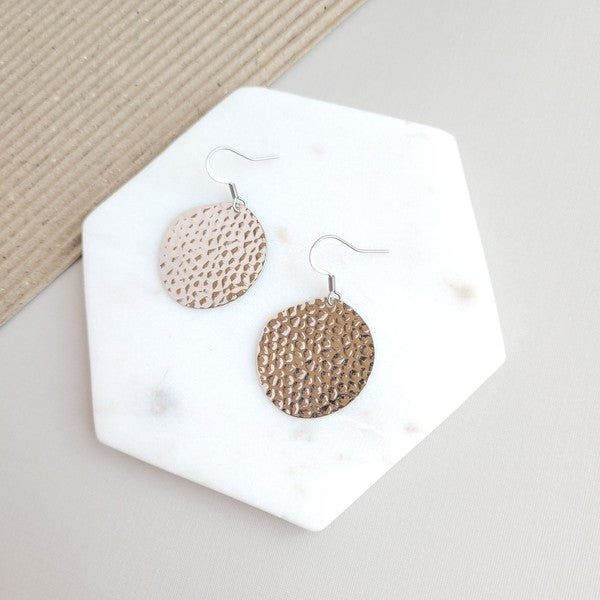 The Lucia Earrings - Silver feature round textured metal designs with stainless steel hooks, arranged on a white hexagonal marble surface at a diagonal angle, set against a subtle beige background. These elegant accessories are crafted with silver-plated hammered brass, offering a timeless appeal as part of the Lucia collection.