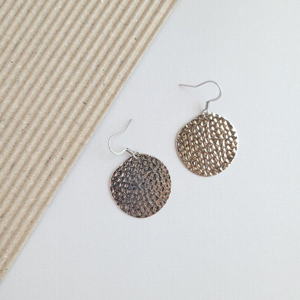 The Lucia Earrings - Silver feature round textured metal designs with stainless steel hooks, arranged on a white hexagonal marble surface at a diagonal angle, set against a subtle beige background. These elegant accessories are crafted with silver-plated hammered brass, offering a timeless appeal as part of the Lucia collection.