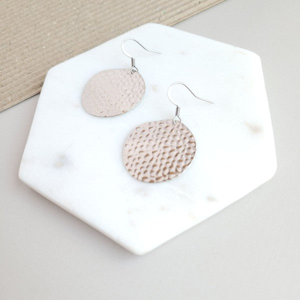 The Lucia Earrings - Silver feature round textured metal designs with stainless steel hooks, arranged on a white hexagonal marble surface at a diagonal angle, set against a subtle beige background. These elegant accessories are crafted with silver-plated hammered brass, offering a timeless appeal as part of the Lucia collection.