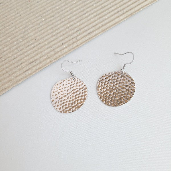 The Lucia Earrings - Silver feature round textured metal designs with stainless steel hooks, arranged on a white hexagonal marble surface at a diagonal angle, set against a subtle beige background. These elegant accessories are crafted with silver-plated hammered brass, offering a timeless appeal as part of the Lucia collection.