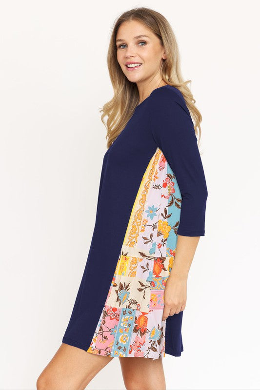 A woman in a Side Contrast A Line Tunic Dress with floral side panels and three-quarter sleeves stands against a white background, smiling and looking to the right. The black dress, made of polyester spandex, complements her black heeled sandals.
