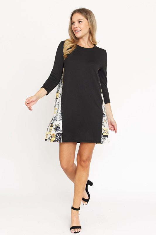 A woman in a Side Contrast A Line Tunic Dress with floral side panels and three-quarter sleeves stands against a white background, smiling and looking to the right. The black dress, made of polyester spandex, complements her black heeled sandals.