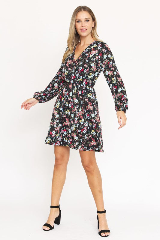 A woman stands against a white paneled wall, wearing the V-Neck Flowy Floral Mini Dress in a maroon floral pattern. The dress features long sleeves and falls gracefully above the knee. She is smiling with one hand on her hip. Made from 95% polyester and 5% spandex, this elegant dress is proudly crafted in the United States.