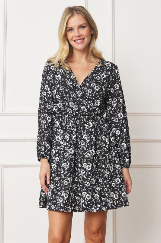 A person with long blonde hair, wearing a black V-Neck Flowy Floral Mini Dress, stands against a white wall, smiling.