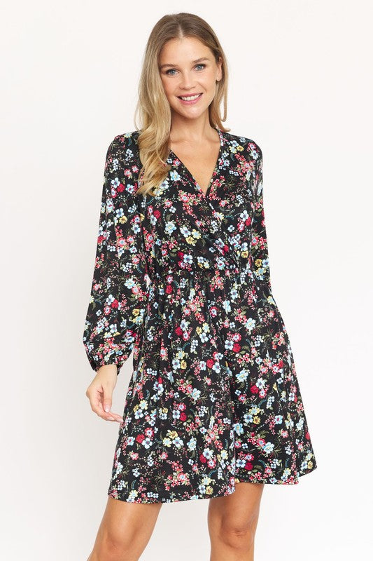 A person with long blonde hair, wearing a black V-Neck Flowy Floral Mini Dress, stands against a white wall, smiling.