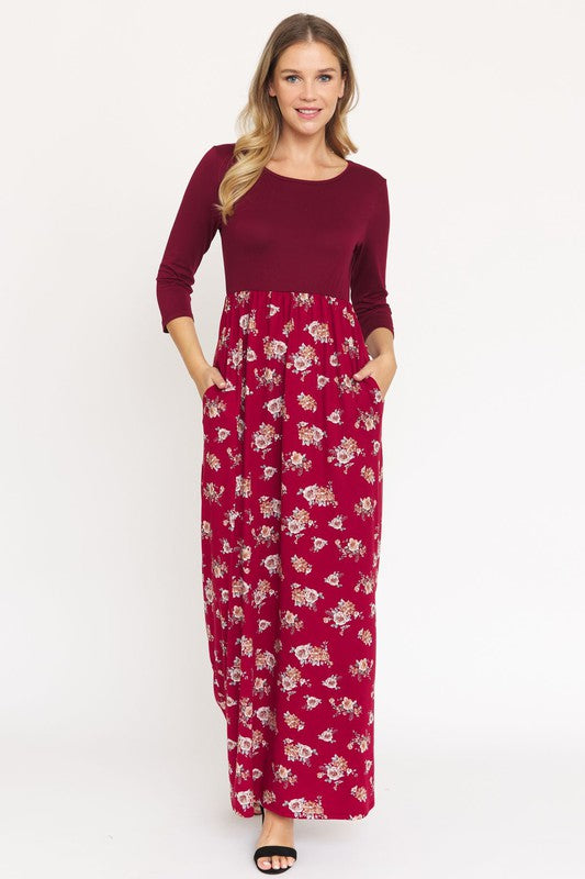 A person is wearing the Quarter Sleeve Floral Maxi Dress in burgundy, featuring a fitted top and a flowing skirt adorned with a delicate floral pattern. This dress, made from a blend of polyester and spandex, has convenient pockets. They have paired it with black sandals.