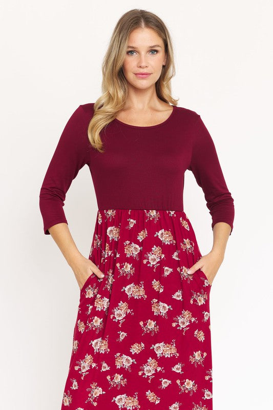 A person is wearing the Quarter Sleeve Floral Maxi Dress in burgundy, featuring a fitted top and a flowing skirt adorned with a delicate floral pattern. This dress, made from a blend of polyester and spandex, has convenient pockets. They have paired it with black sandals.