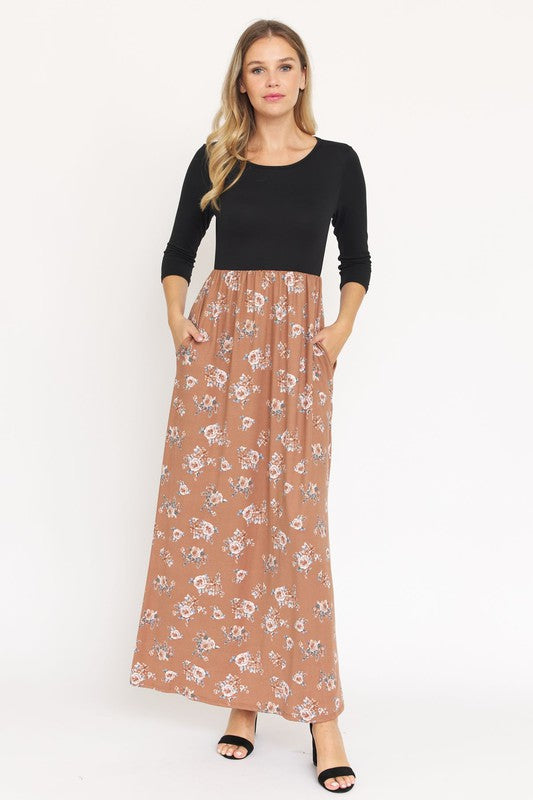 A woman stands against a plain background, wearing the Quarter Sleeve Floral Maxi Dress, which features a black top and a floral-patterned brown skirt. Her hands are in her pockets, and she is also wearing black sandals.