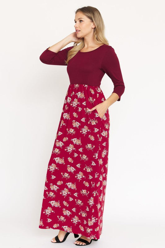 A person is wearing the Quarter Sleeve Floral Maxi Dress in burgundy, featuring a fitted top and a flowing skirt adorned with a delicate floral pattern. This dress, made from a blend of polyester and spandex, has convenient pockets. They have paired it with black sandals.