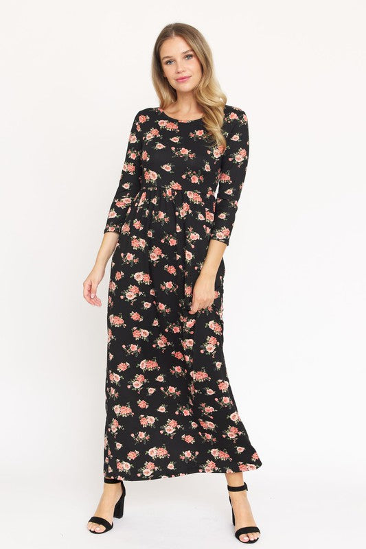 A woman in the Bouquet Floral Quarter Sleeve Maxi Dress poses against a plain white background. She is standing with one hand touching her hair and wearing black heels, showcasing the elegance of her U.S.-made black floral maxi dress.