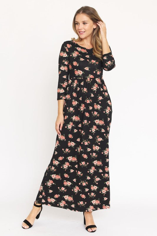 A woman in the Bouquet Floral Quarter Sleeve Maxi Dress poses against a plain white background. She is standing with one hand touching her hair and wearing black heels, showcasing the elegance of her U.S.-made black floral maxi dress.