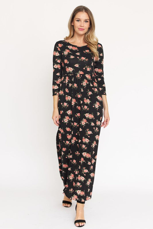 A woman in the Bouquet Floral Quarter Sleeve Maxi Dress poses against a plain white background. She is standing with one hand touching her hair and wearing black heels, showcasing the elegance of her U.S.-made black floral maxi dress.