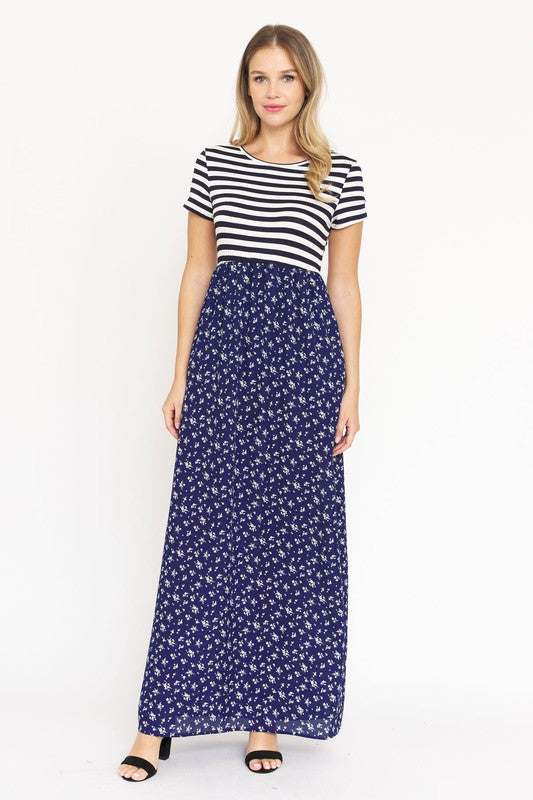 A woman is wearing a black and white striped top paired with the Short Sleeve Floral Maxi Dress in light blue. She is standing against a plain white background, showcasing the effortless beauty of this made-in-the-USA ensemble.