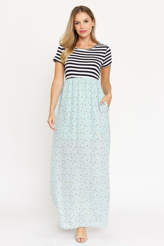 A woman is wearing a black and white striped top paired with the Short Sleeve Floral Maxi Dress in light blue. She is standing against a plain white background, showcasing the effortless beauty of this made-in-the-USA ensemble.