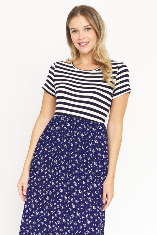 A woman is wearing a black and white striped top paired with the Short Sleeve Floral Maxi Dress in light blue. She is standing against a plain white background, showcasing the effortless beauty of this made-in-the-USA ensemble.