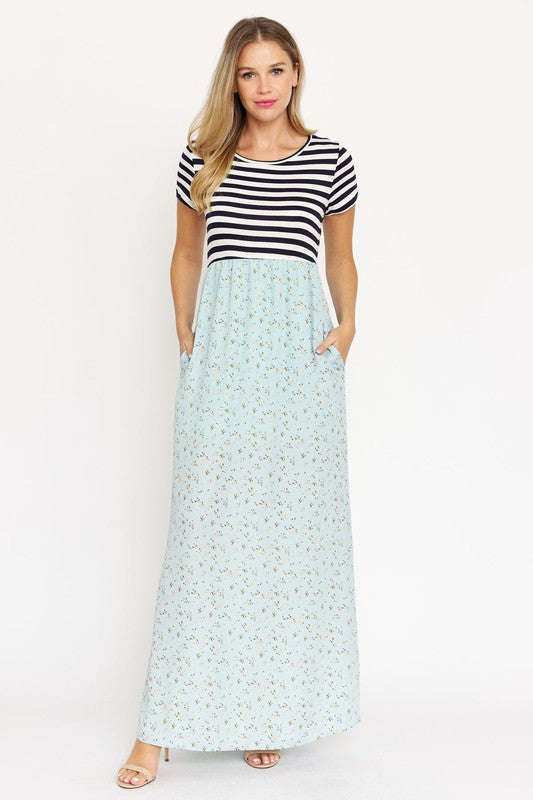 A woman is wearing a black and white striped top paired with the Short Sleeve Floral Maxi Dress in light blue. She is standing against a plain white background, showcasing the effortless beauty of this made-in-the-USA ensemble.