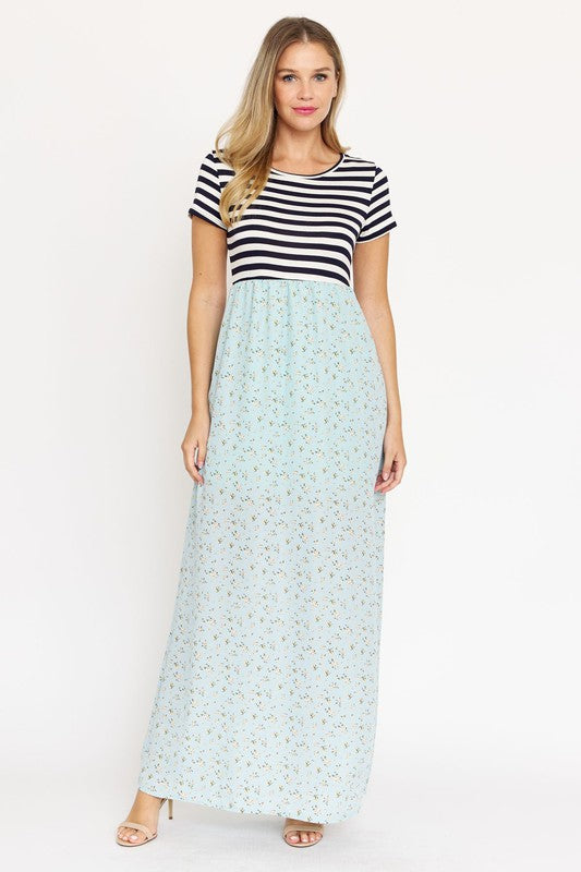 A woman is wearing a black and white striped top paired with the Short Sleeve Floral Maxi Dress in light blue. She is standing against a plain white background, showcasing the effortless beauty of this made-in-the-USA ensemble.