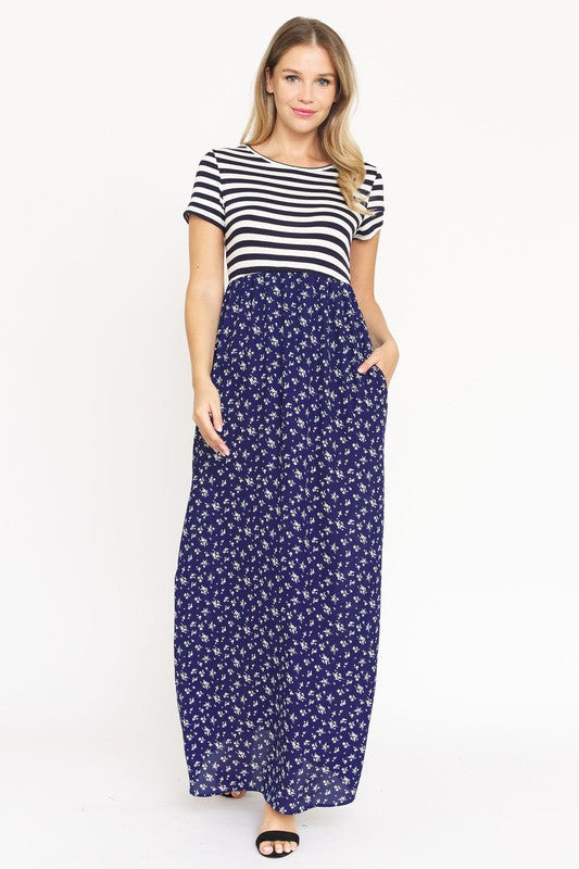 A woman is wearing a black and white striped top paired with the Short Sleeve Floral Maxi Dress in light blue. She is standing against a plain white background, showcasing the effortless beauty of this made-in-the-USA ensemble.