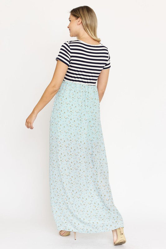 A woman is wearing a black and white striped top paired with the Short Sleeve Floral Maxi Dress in light blue. She is standing against a plain white background, showcasing the effortless beauty of this made-in-the-USA ensemble.