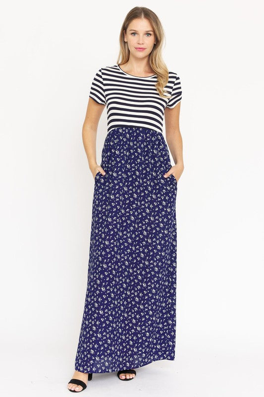 A woman is wearing a black and white striped top paired with the Short Sleeve Floral Maxi Dress in light blue. She is standing against a plain white background, showcasing the effortless beauty of this made-in-the-USA ensemble.