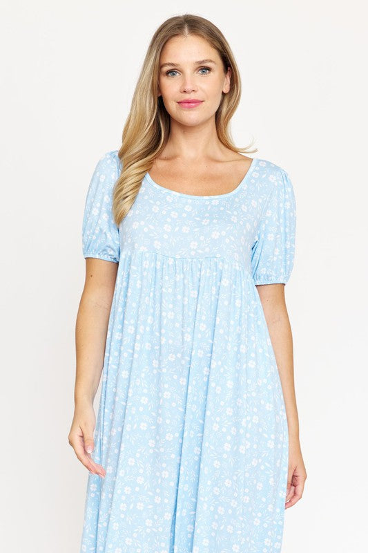 A woman is wearing the Empire Waist Floral Puff Sleeve Dress, which is light pink with an ankle-length cut and features a floral pattern. The dress includes short, floral puff sleeves. She completes her outfit with white sneakers and stands against a plain white background.