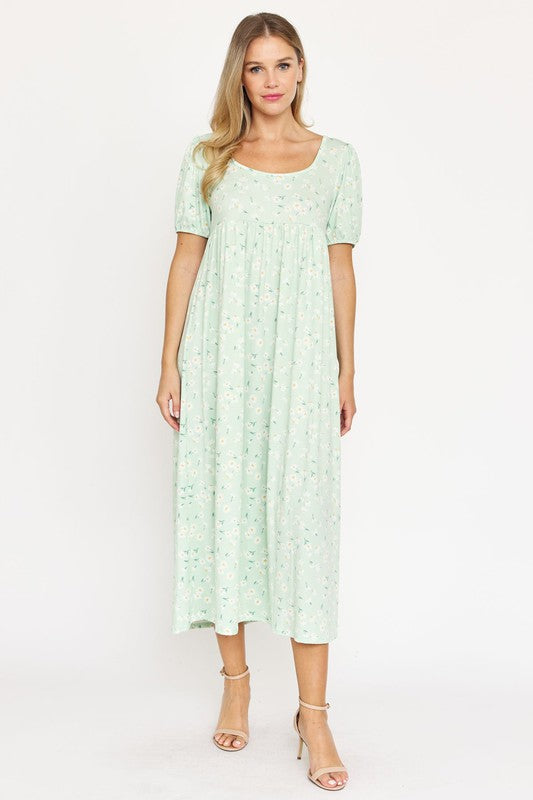 A woman is wearing the Empire Waist Floral Puff Sleeve Dress, which is light pink with an ankle-length cut and features a floral pattern. The dress includes short, floral puff sleeves. She completes her outfit with white sneakers and stands against a plain white background.