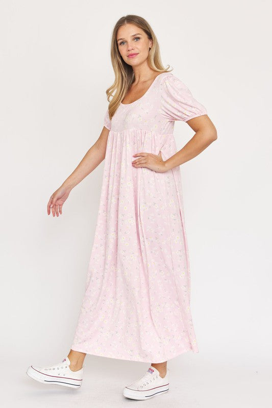 A woman is wearing the Empire Waist Floral Puff Sleeve Dress, which is light pink with an ankle-length cut and features a floral pattern. The dress includes short, floral puff sleeves. She completes her outfit with white sneakers and stands against a plain white background.