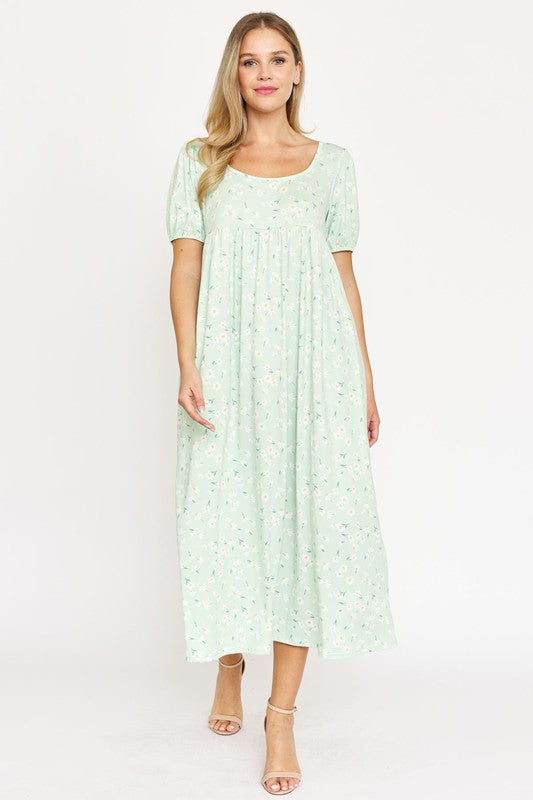 A woman is wearing the Empire Waist Floral Puff Sleeve Dress, which is light pink with an ankle-length cut and features a floral pattern. The dress includes short, floral puff sleeves. She completes her outfit with white sneakers and stands against a plain white background.