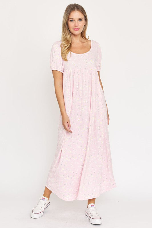 A woman is wearing the Empire Waist Floral Puff Sleeve Dress, which is light pink with an ankle-length cut and features a floral pattern. The dress includes short, floral puff sleeves. She completes her outfit with white sneakers and stands against a plain white background.