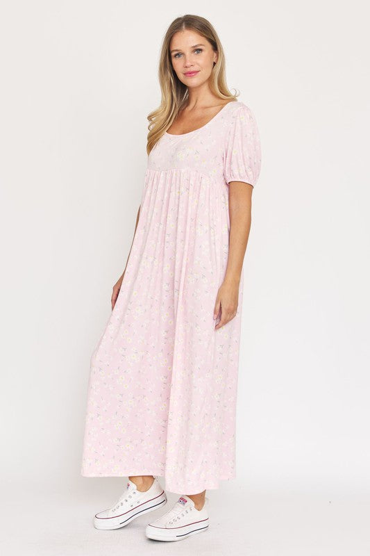 A woman is wearing the Empire Waist Floral Puff Sleeve Dress, which is light pink with an ankle-length cut and features a floral pattern. The dress includes short, floral puff sleeves. She completes her outfit with white sneakers and stands against a plain white background.