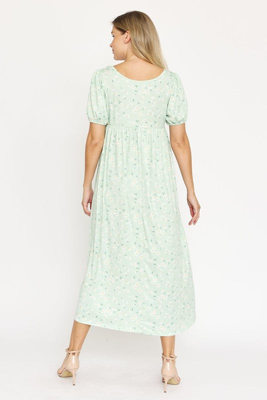 A woman is wearing the Empire Waist Floral Puff Sleeve Dress, which is light pink with an ankle-length cut and features a floral pattern. The dress includes short, floral puff sleeves. She completes her outfit with white sneakers and stands against a plain white background.