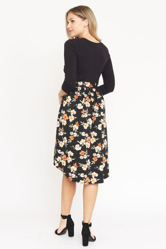 A woman in a Surplice Floral Sash Midi Dress, comprised of 95% Polyester and 5% Spandex, stands against a plain white backdrop. The knee-length dress, designed in navy blue with a floral skirt and wrapped bodice, showcases elegant craftsmanship from the United States.