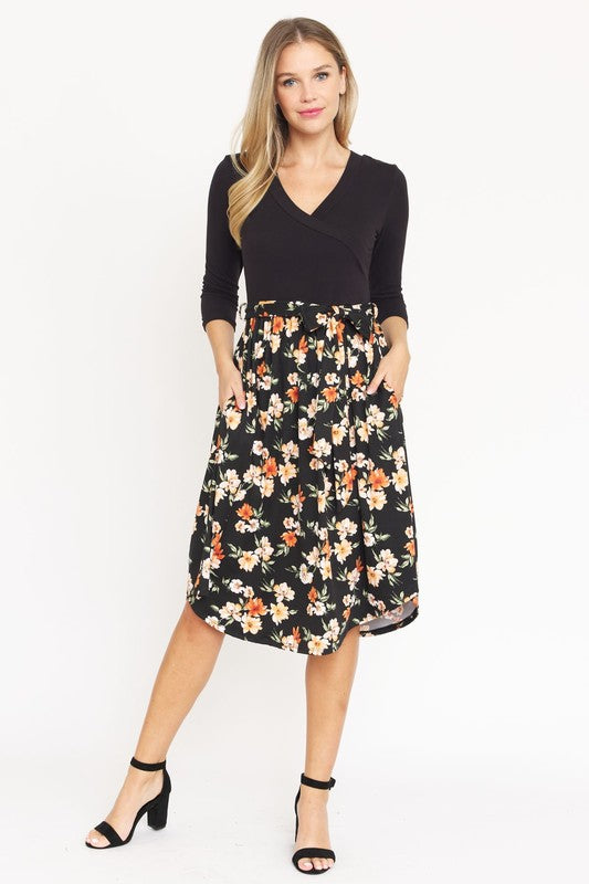 A woman in a Surplice Floral Sash Midi Dress, comprised of 95% Polyester and 5% Spandex, stands against a plain white backdrop. The knee-length dress, designed in navy blue with a floral skirt and wrapped bodice, showcases elegant craftsmanship from the United States.