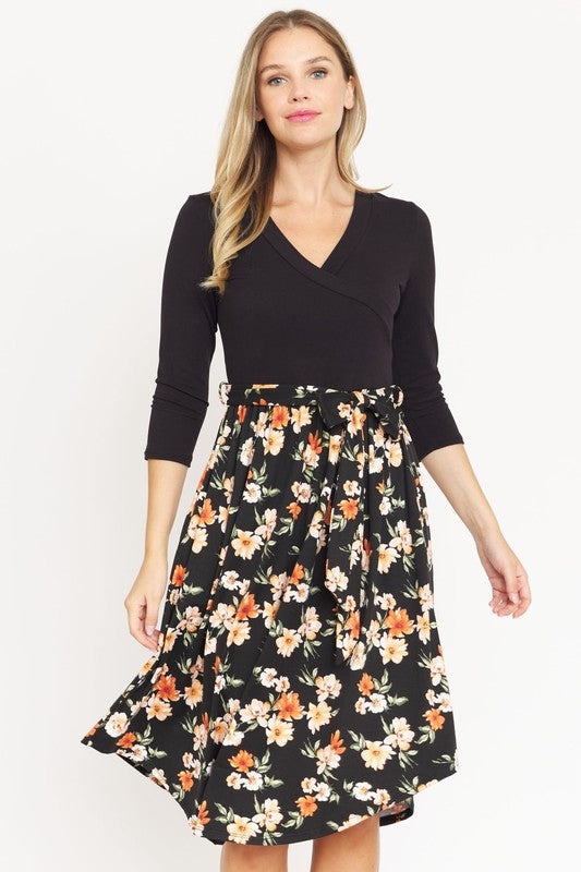 A woman in a three-quarter sleeve black top and a Surplice Floral Sash Midi Dress waves, smiling.