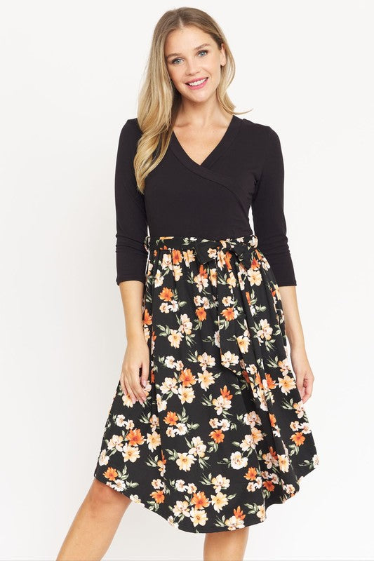 A woman in a Surplice Floral Sash Midi Dress, comprised of 95% Polyester and 5% Spandex, stands against a plain white backdrop. The knee-length dress, designed in navy blue with a floral skirt and wrapped bodice, showcases elegant craftsmanship from the United States.