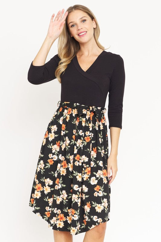 A woman in a three-quarter sleeve black top and a Surplice Floral Sash Midi Dress waves, smiling.